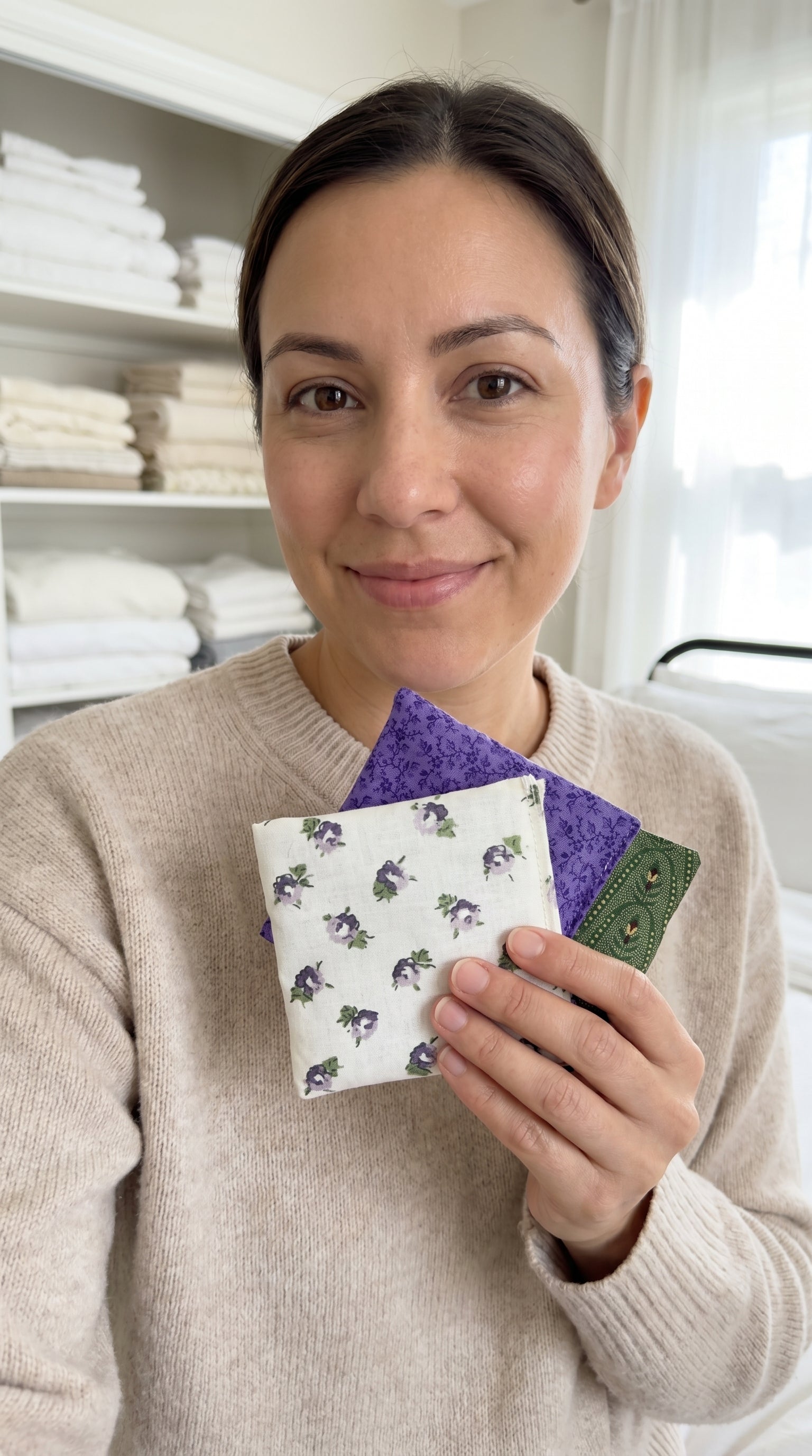 Woman holding 3 lavender sachets in a  floral pattern in a room with shelves of folded towels.